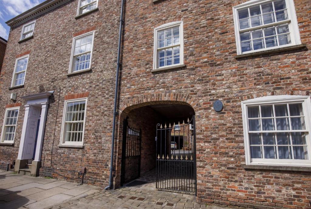 A brick building with white sash windows and a large arched gateway with an ornate black iron gate. The entrance leads to a courtyard. A blue plaque is mounted on the wall near the second window from the left.