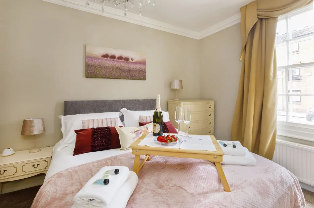 A cosy bedroom with a large bed adorned with red and pink pillows and a tray holding a champagne bottle, glasses, and strawberries. A chandelier hangs from the ceiling, and a window with gold curtains brightens the room. A vanity table is nearby.