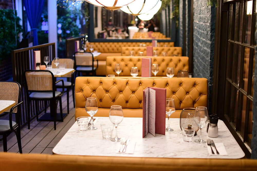 A cosy restaurant with yellow tufted booth seating, marble tables set with wine glasses, menus, and utensils. Warm lighting creates an inviting atmosphere, with chairs and decorative lanterns enhancing the elegant ambiance.