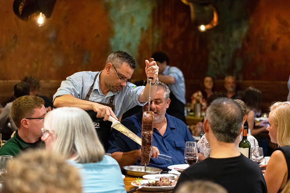 A server in a restaurant carves meat from a skewer onto a diners plate. The setting is busy, with people seated around tables enjoying their meals. Dim lighting creates a warm, cosy atmosphere. Wine glasses and bottles are visible on the tables.