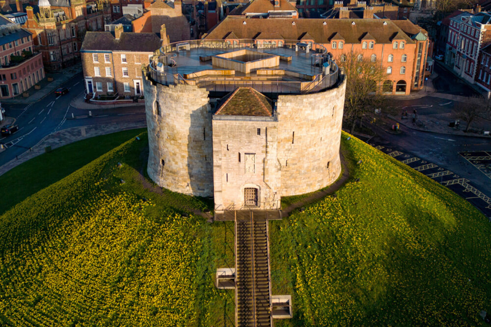 A circular stone castle with a wooden walkway sits atop a grassy hill covered in yellow flowers. Surrounded by historic buildings, the scene captures a blend of medieval architecture and modern urban landscape in an aerial view.