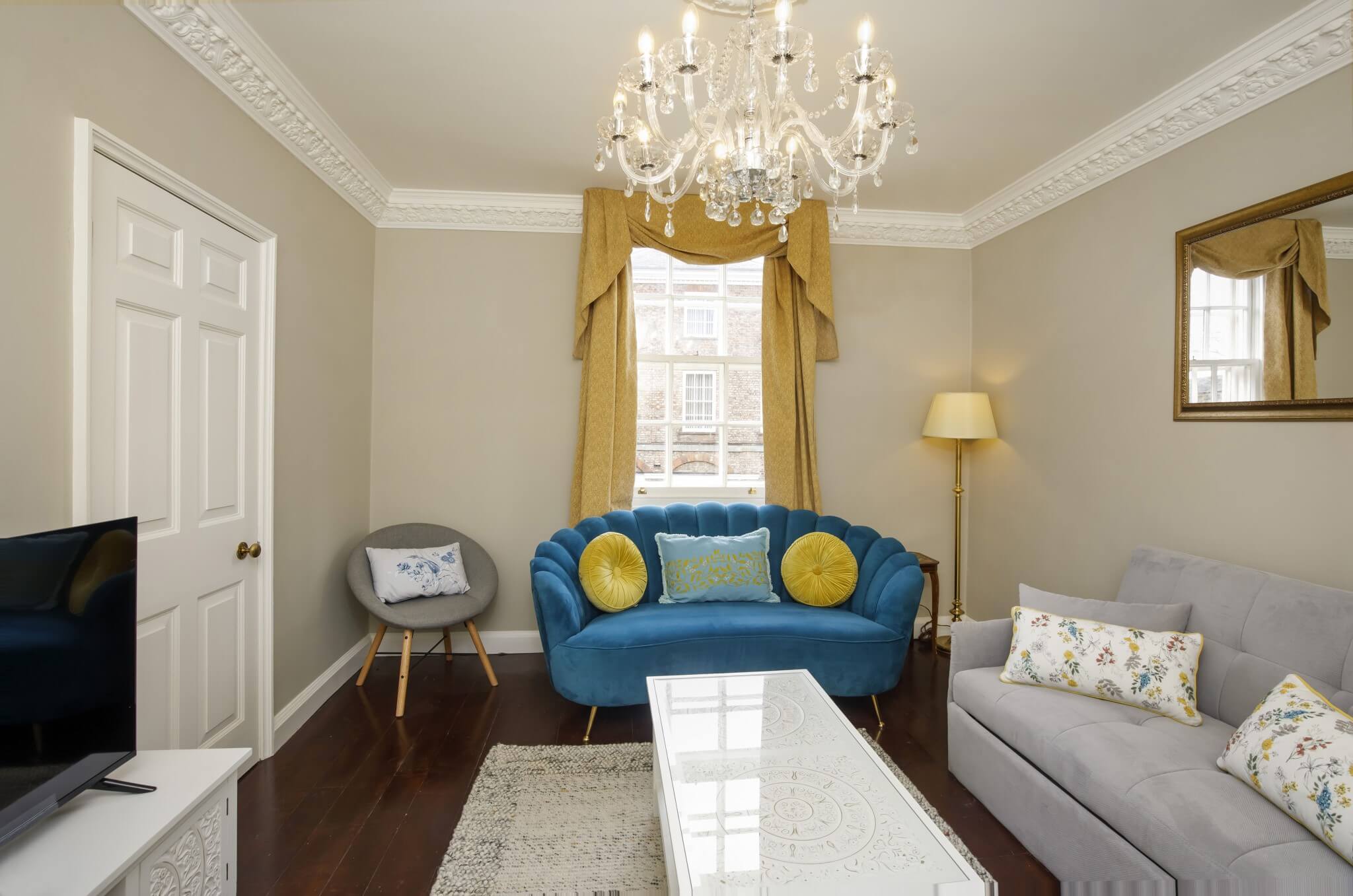 Elegant living room with a blue sofa, grey armchair, and a light grey couch. A chandelier hangs from the ceiling, and there's a large window with golden curtains. A TV and mirror are also visible.