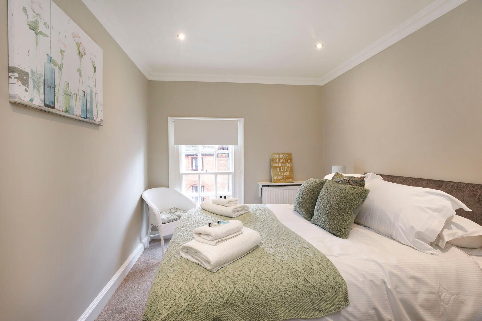 A cosy bedroom with a neatly made bed featuring green pillows and a textured blanket. Towels are folded on the bed. A window lets in natural light. A chair is placed in the corner, and a decorative wall art and sign add charm.