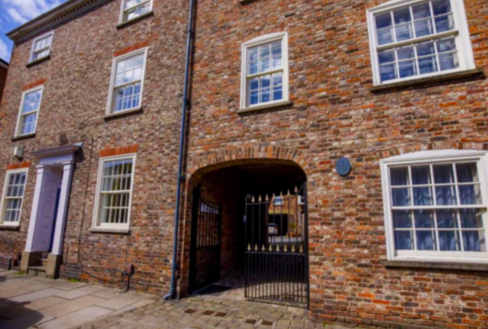 A brick building with white sash windows and a large arched gateway with an ornate black iron gate. The entrance leads to a courtyard. A blue plaque is mounted on the wall near the second window from the left.