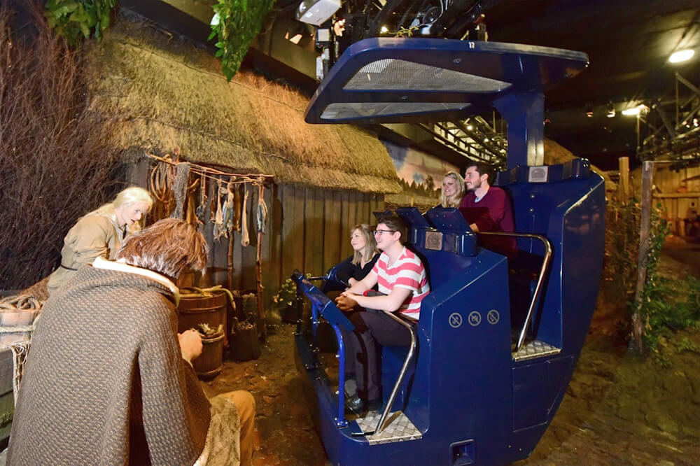 Visitors ride a blue vehicle in an immersive historical exhibit, observing figures in period clothing. The setting appears to be a rustic, old-world village scene with thatched roofs and foliage.