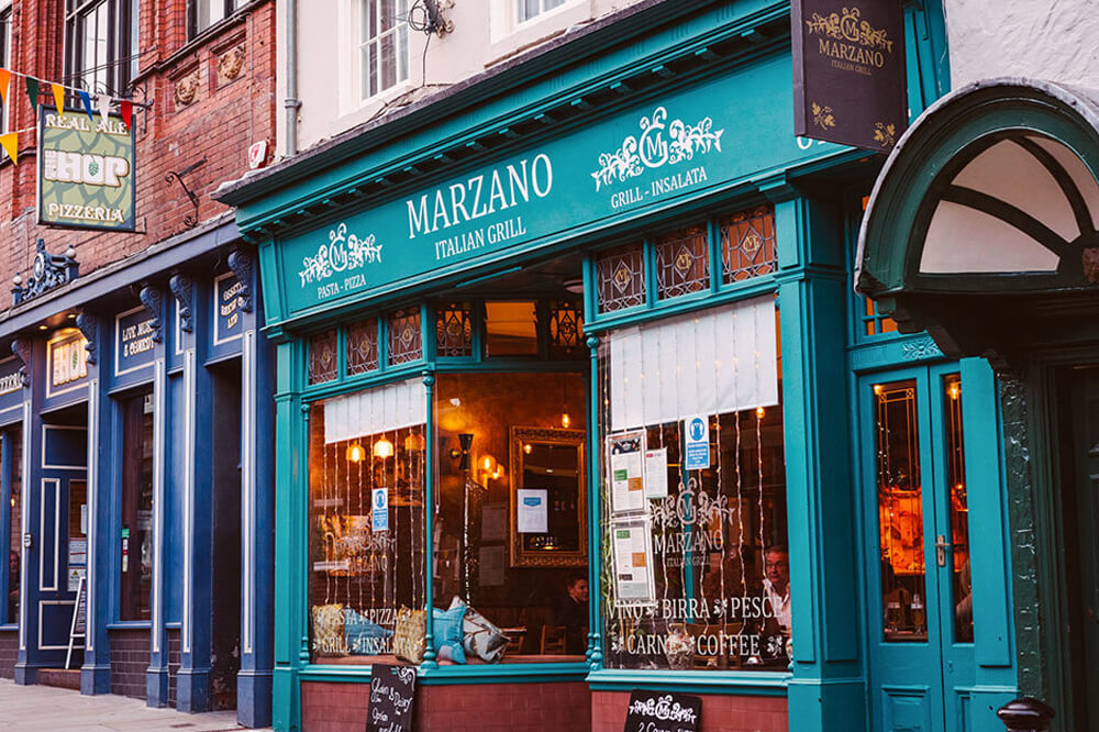 Street view of a teal-painted Italian restaurant called Marzano, featuring signs for pizza, insalata, and grill items. Warm interior lights and customers can be seen through the large windows. Banners hang from neighbouring red brick buildings.