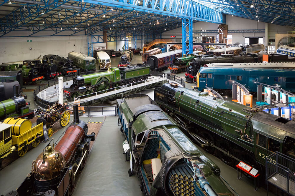 A large indoor museum exhibit featuring various historical trains and locomotives displayed on tracks. The room has a blue steel roof, and some trains are green and blue, with others in black and yellow, all surrounded by walkways for viewing.