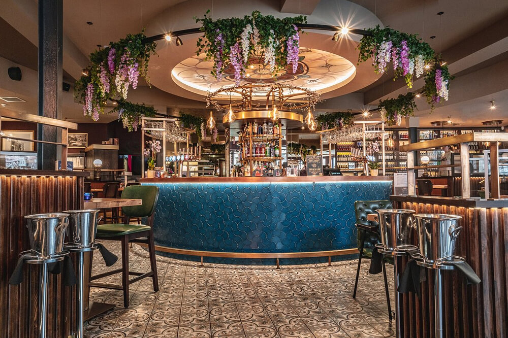 A modern, stylish bar interior featuring a circular blue-tiled bar counter under an ornate ceiling. Hanging greenery and purple flowers add elegance. The floor is tiled, and there are high chairs and tables around the space.