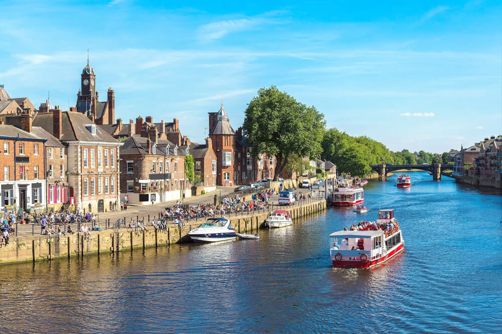 A scenic view of a river with boats docked along its banks, a bridge spanning across, and historic buildings in the background. Lush green trees line the walkway, and the sky is partly cloudy, enhancing the picturesque setting.