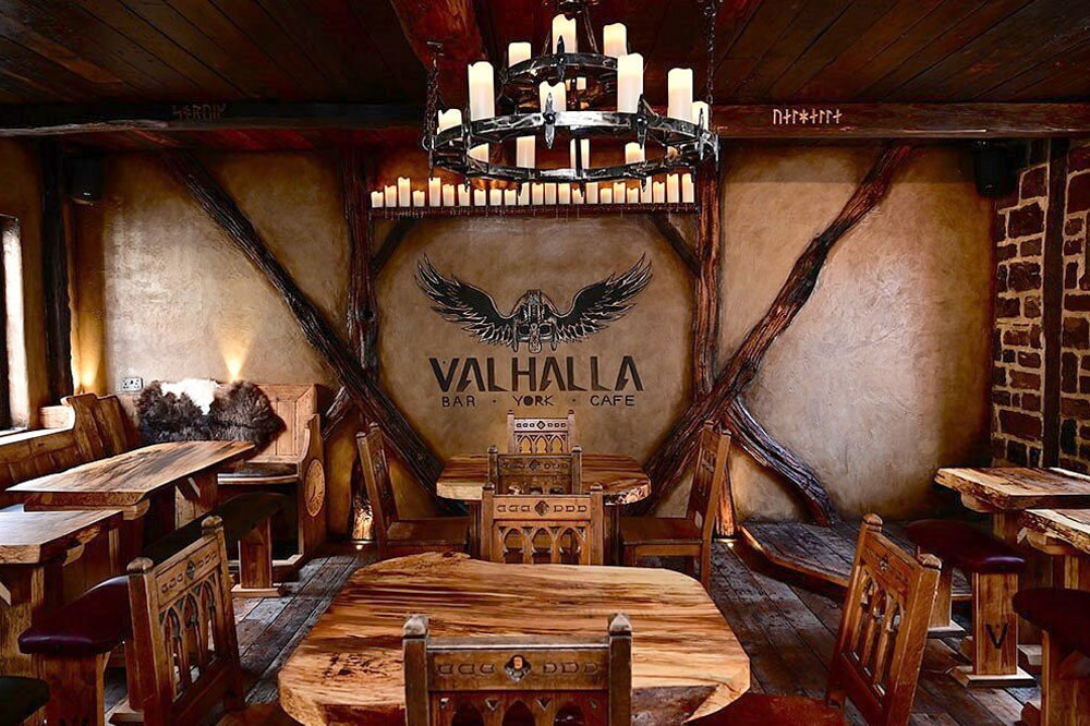 Rustic interior of Valhalla Bar York Café featuring wooden tables and chairs, Viking-themed décor with a winged emblem on the wall, and a chandelier with candles hanging from the ceiling. The walls have exposed beams and Viking runes.