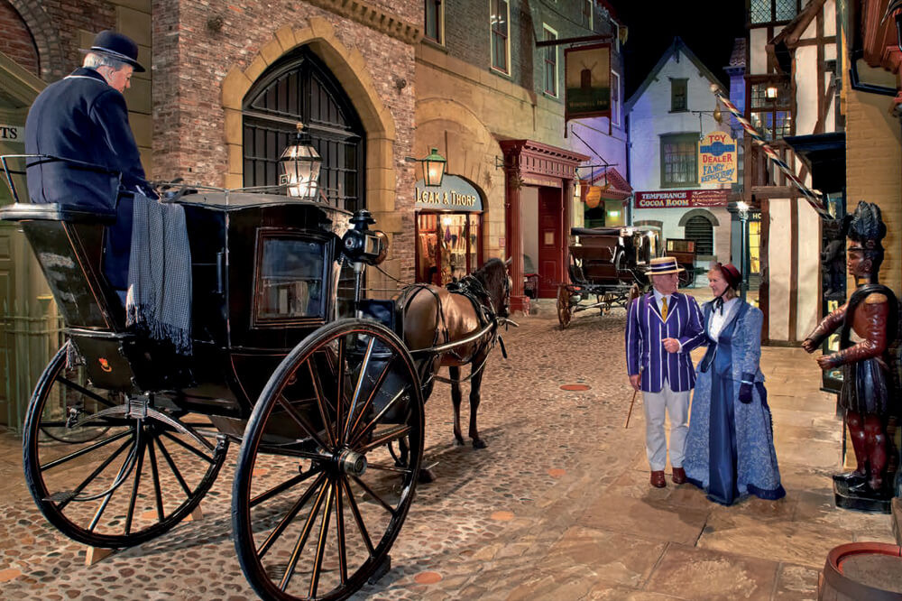 A cobblestone street scene with a horse-drawn carriage. A coachman sits on the carriage, while a man and woman in period attire converse nearby. The street is lined with old-fashioned shops and a lamp post.