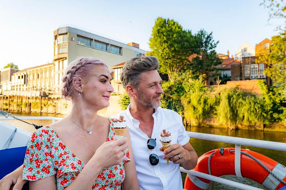 A couple enjoys ice cream cones on a sunny day aboard a boat, with buildings and lush trees lining the riverbank in the background. The woman wears a floral dress, and the man, in a white shirt, looks relaxed. An orange lifebuoy is visible on the boat.