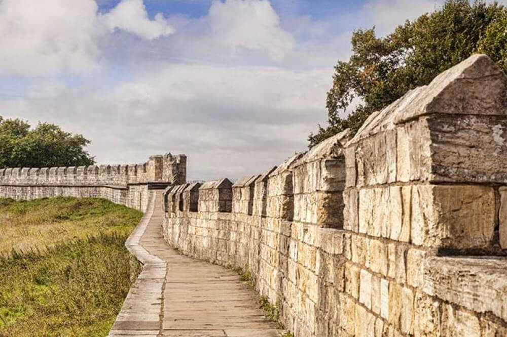 A stone pathway runs along the top of a historic wall, bordered by grass and trees under a partly cloudy sky. The wall features crenelations and extends into the distance.