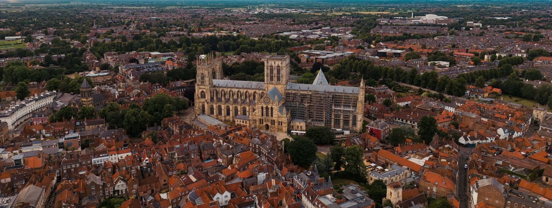 Aerial view of a large, historic cathedral surrounded by a dense array of red-roofed buildings and lush green trees. The landscape extends to a distant horizon under a clear blue sky.