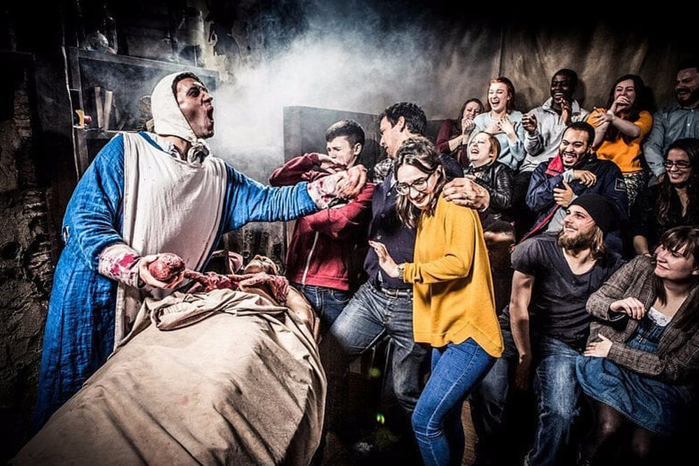 A group of people, some smiling and others looking startled, watch a dramatic actor in medieval costume performing in a dimly lit room with fog. A person lies covered on a table nearby. The setting has a tense, theatrical atmosphere.