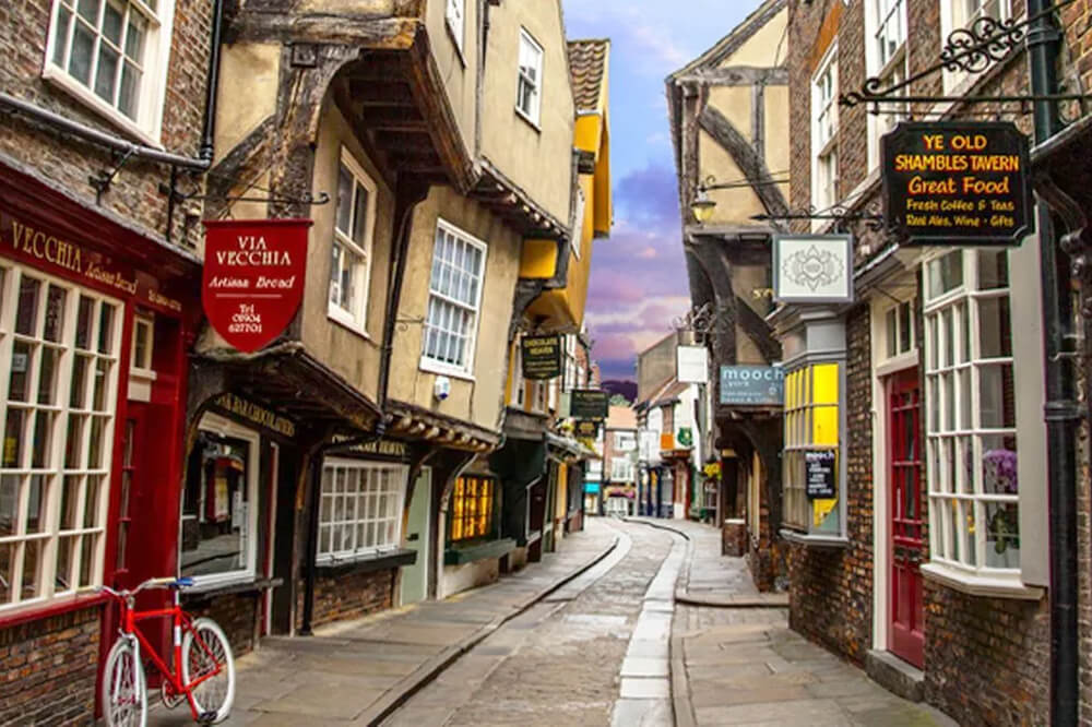 A narrow, cobbled street lined with historic, leaning buildings featuring colourful shop signs. A red bicycle is parked outside a window with red frames. The sky is slightly overcast, adding a quaint charm to the scene.