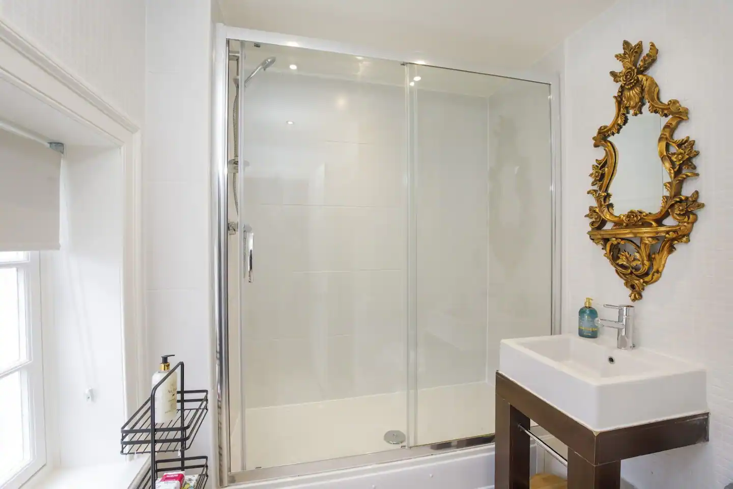 A modern bathroom with a glass-enclosed shower, a white sink on a dark wooden stand, and a decorative gold-framed mirror. There is a window with a white blind and a shelf holding toiletries beside the shower.