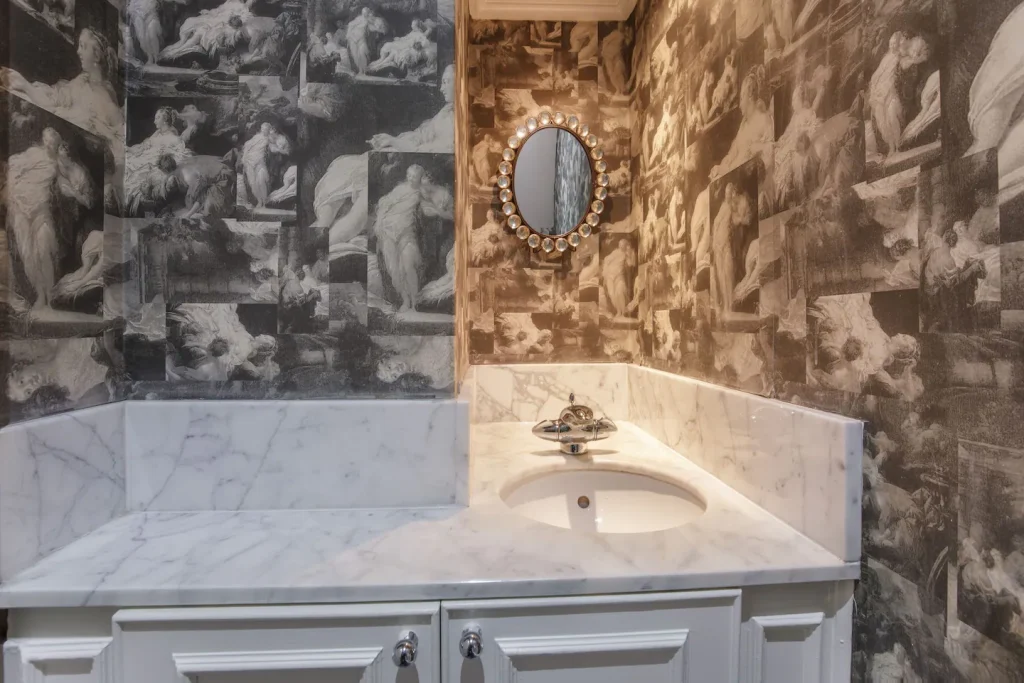 A small bathroom with a white marble countertop and sink. The walls are adorned with black and white classical art-themed wallpaper. A decorative round mirror with a detailed frame hangs above the sink, reflecting part of the patterned wallpaper.