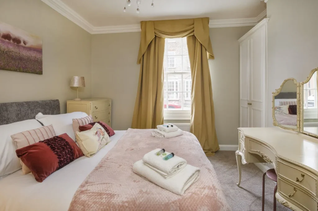 A cosy bedroom with a large bed featuring plush pillows and a pink blanket. A window with curtains overlooks the street. A dresser and a dressing table with a mirror are on the right, and a soft light fills the room.