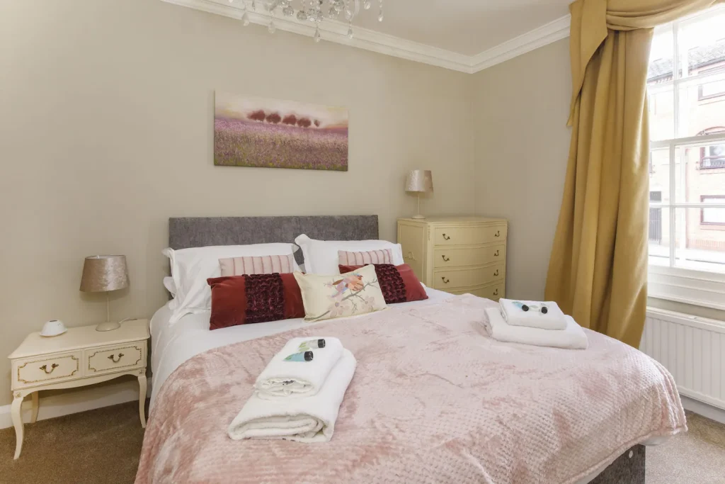 A cozy bedroom with a double bed featuring pink and white bedding and accent pillows. Towels are neatly placed at the foot of the bed. The room is decorated with elegant furniture, two bedside tables, a dresser, and a large window with gold curtains.