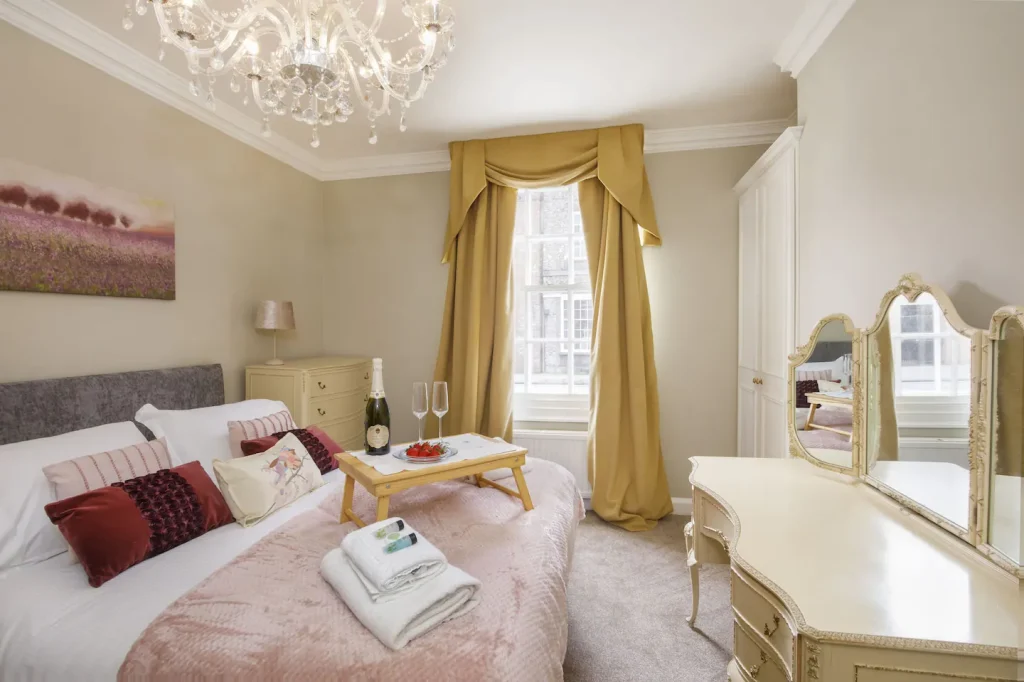 A cosy bedroom with a large bed adorned with red and pink pillows and a tray holding a champagne bottle, glasses, and strawberries. A chandelier hangs from the ceiling, and a window with gold curtains brightens the room. A vanity table is nearby.