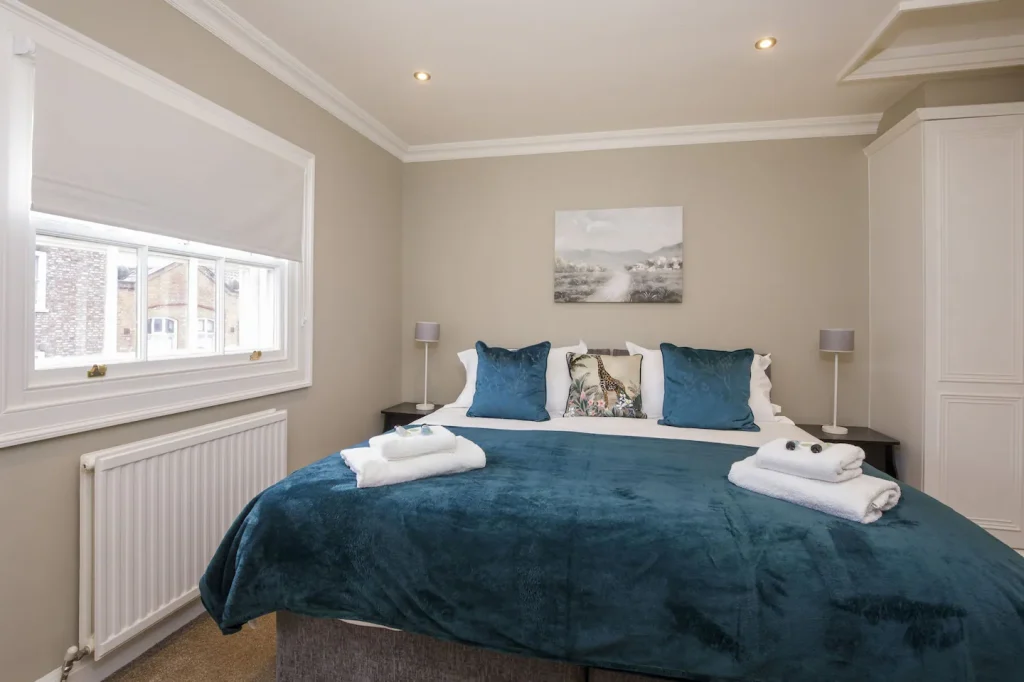 A cosy bedroom featuring a large bed with a blue throw and patterned pillows, flanked by nightstands with lamps. A landscape painting hangs above the bed. Natural light enters through a window with a white blind. Towels are neatly arranged on the bed.