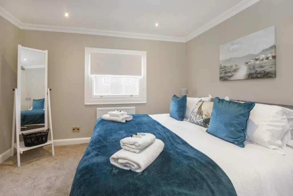 A cosy bedroom with a large bed featuring blue and white bedding and two neatly folded towels. A tall mirror stands beside the window with a closed blind. A landscape painting hangs on the light grey wall, and the carpet is beige.