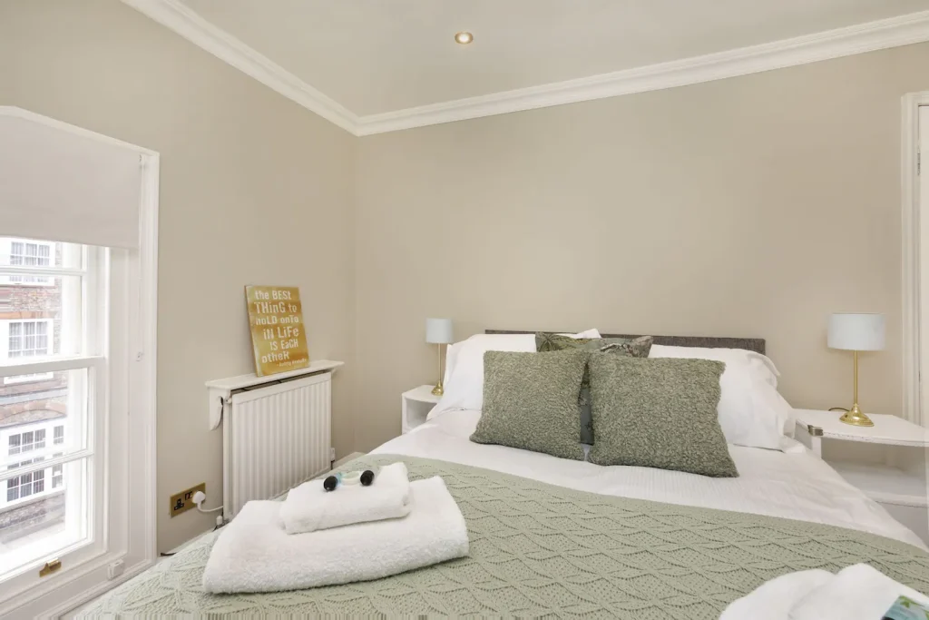 A cosy bedroom with a neatly made bed featuring green and white bedding, two green pillows, and folded towels on top. The room has two small bedside tables with lamps, a radiator, and a sign on the wall near a window with a street view.