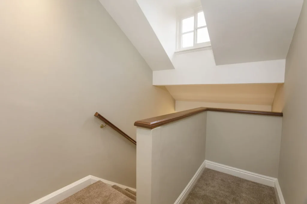 A staircase with beige carpet and light grey walls leads up to a landing. A wooden handrail lines the left side. Sunlight filters through a small, rectangular window above, illuminating the space.