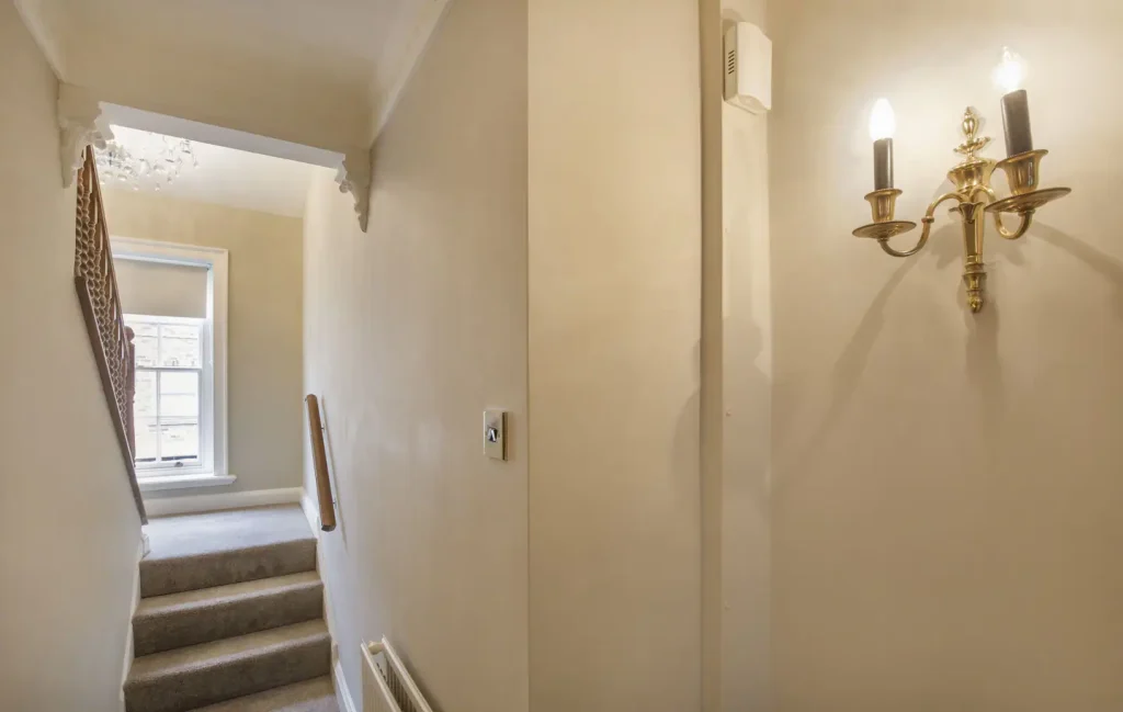 A well-lit hallway featuring a carpeted staircase leading to a window. The cream-colored walls are adorned with a classic two-bulb wall sconce. Natural light filters through the window, casting soft shadows on the wall.