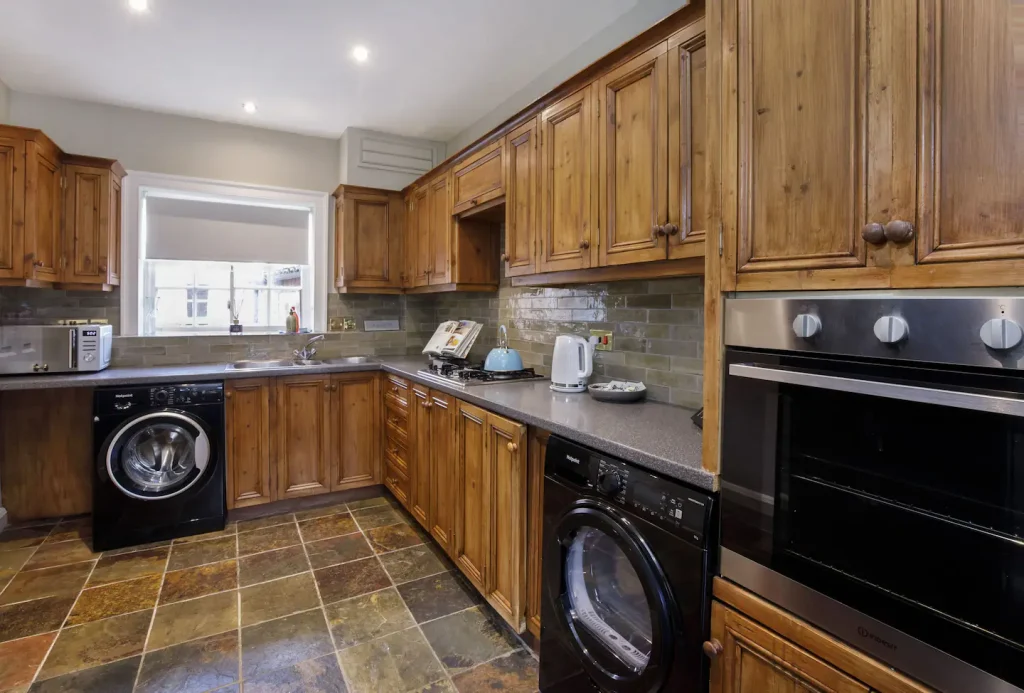 A kitchen with wooden cabinets, stone tile flooring, and a dual oven. It has a microwave, a washing machine, a dryer, and a kettle on the countertop. A wide window is above the sink, letting in natural light.