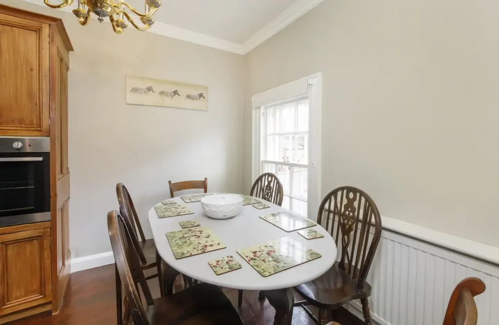 A dining room with an oval table set for six, featuring floral placemats and a white bowl centerpiece. Wooden chairs surround the table. A wooden cabinet and an oven are on the left. A window and a horse picture adorn the light-coloured walls.