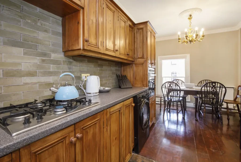 A kitchen with wooden cabinets and a countertop holding a blue kettle and white toaster. A gas stove is built into the counter. The dining area has a wooden table with chairs, and a chandelier hangs overhead.