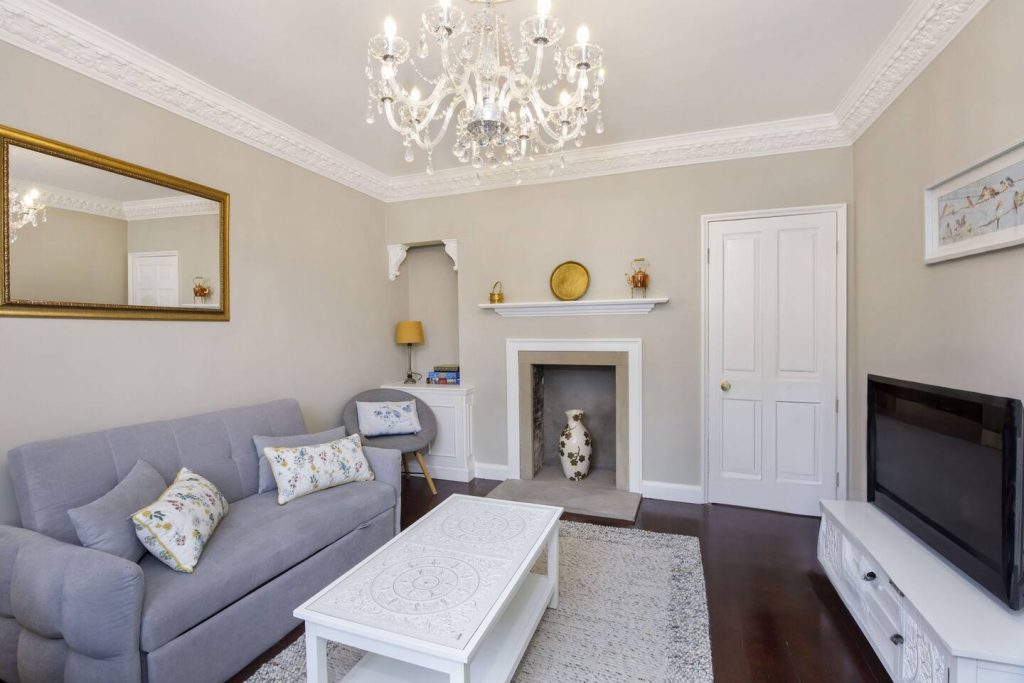 A living room with a grey sofa, decorative pillows, white coffee table, and a chandelier. The room has a fireplace, mirror, and a TV on a stand. Walls are light grey with white trim.