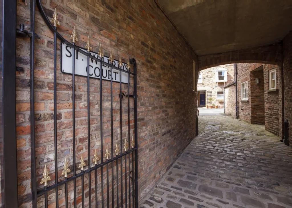 A cobblestone alleyway leads to a brick courtyard. A black wrought iron gate with golden fleur-de-lis features and a sign reading St. Michaels Court partially closed on the left, framed by an arched brick entrance.