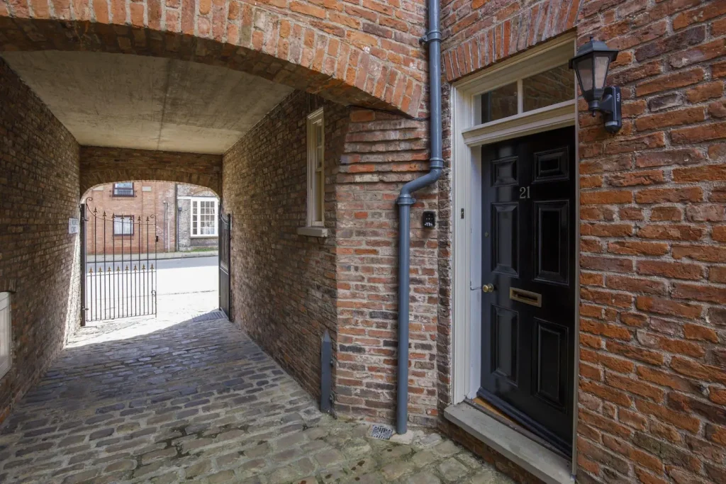 Cobblestone path under an arched brick passageway, leading to a street. A black door with the number 21 is on the right side, set in a brick wall. A metal gate is visible at the end of the passage.